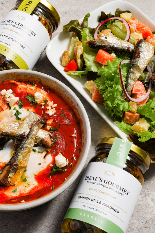 Two dishes with sardines and a bottle of Irene's Gourmet Spanish Style Sardines on a textured surface.